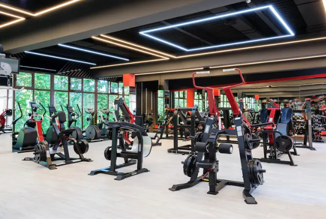 Modern gym interior with various exercise machines and fitness equipment arranged in a spacious, well-lit room with large windows and LED ceiling lights.