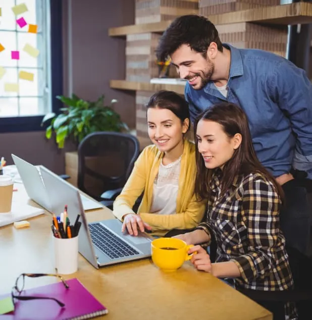 Three coworkers gathered around a laptop at a wooden desk in an office. Two women are seated, one holding a yellow coffee mug, while a man stands behind them, all smiling and looking at the screen. The desk has pens, a notebook, and other office supplies, with a plant and sticky notes visible in the background