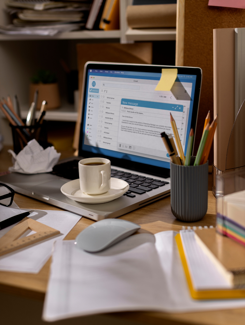 A cluttered desk with a laptop displaying an email inbox and a new message draft. A small cup of coffee on a saucer sits on the laptop keyboard. Surrounding the laptop are pencils in a holder, scattered papers, a triangular ruler, a notebook, and a computer mouse. Shelves with files and office supplies are visible in the background.