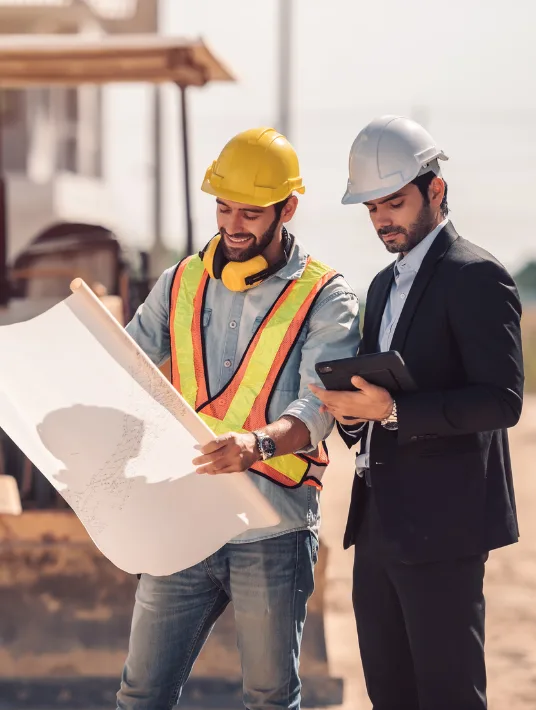 A construction worker in a yellow safety vest and helmet, reviewing plans, while a professional in a suit, also wearing a helmet, checks a tablet on a construction site.