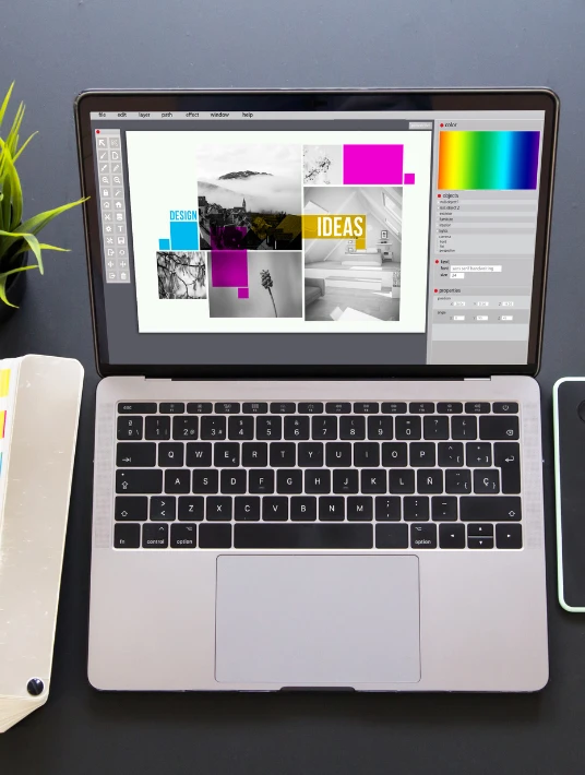 A top view of a laptop showing a graphic design project with colorful blocks and the word 'IDEAS' displayed on the screen. The workspace includes a notepad, a plant, and a smartphone.