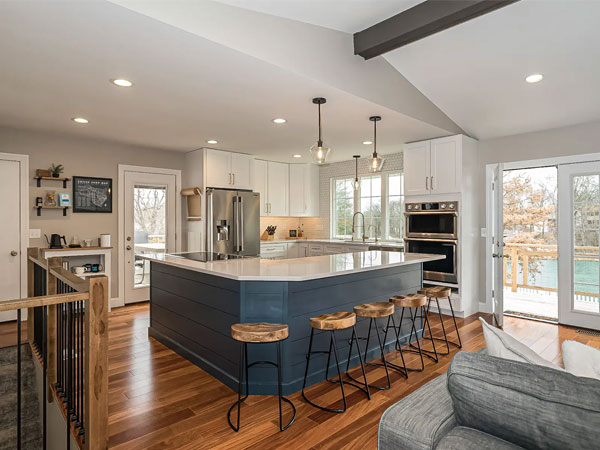 A modern kitchen with a large island, four bar stools, white cabinetry, stainless steel appliances, and natural light streaming through the windows.