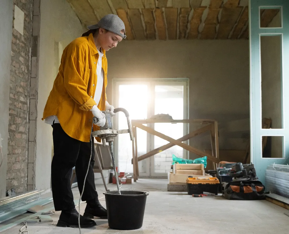 A person in a yellow jacket and cap is mixing materials with an electric mixer in a construction site. The background shows unfinished walls and construction tools