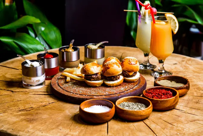 A delicious food spread consisting of three mini burgers placed on a wooden board, surrounded by small bowls containing various spices such as salt, pepper, and chili powder. Two colorful cocktails with garnishes are also visible on the side, adding to the vibrant presentation.