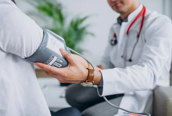 A doctor is measuring the blood pressure of a patient using a blood pressure cuff. The doctor is wearing a white coat with a stethoscope around their neck, while the patient’s arm is exposed as the cuff is placed around their upper arm. The setting appears to be a medical clinic or office with a plant in the background.