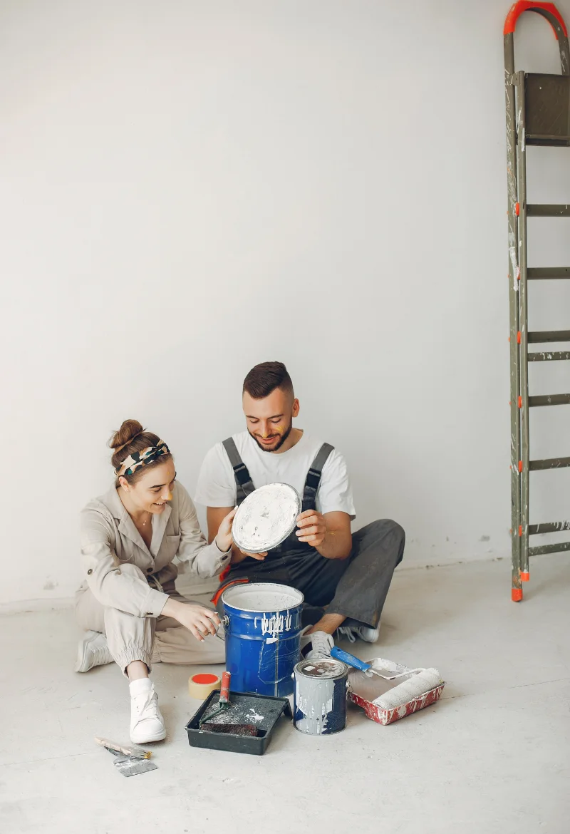 A man and a woman are sitting on the floor, surrounded by paint supplies. They are smiling and holding paint cans, preparing to start painting. A ladder is seen in the background