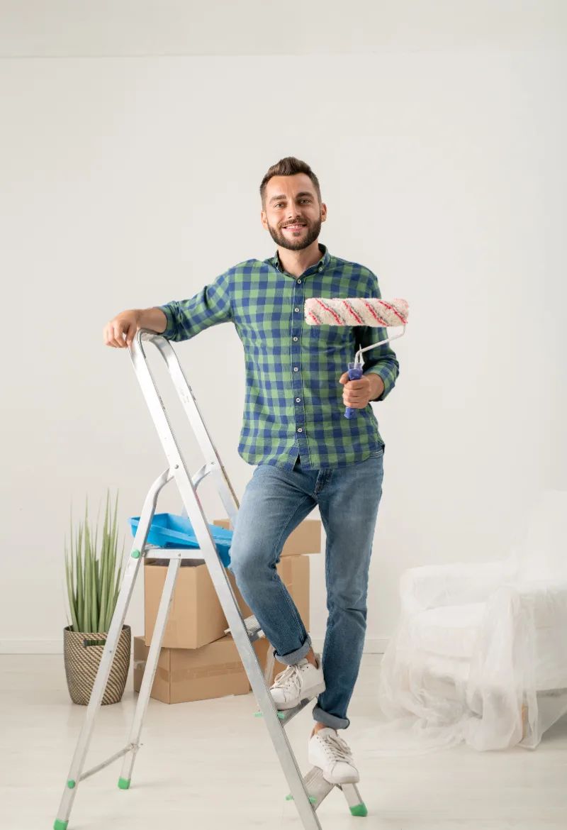 A man smiling while standing on a ladder, holding a paint roller, ready to paint a wall in a room. The setting includes boxes and a houseplant, indicating a home remodel project