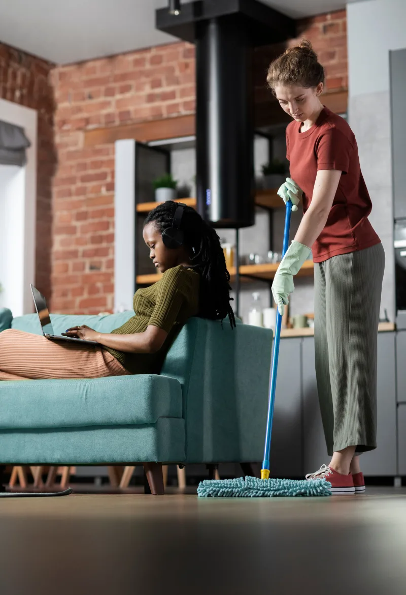 A woman in green rubber gloves is mopping the floor while another woman, wearing headphones, is sitting on a sofa working on a laptop
