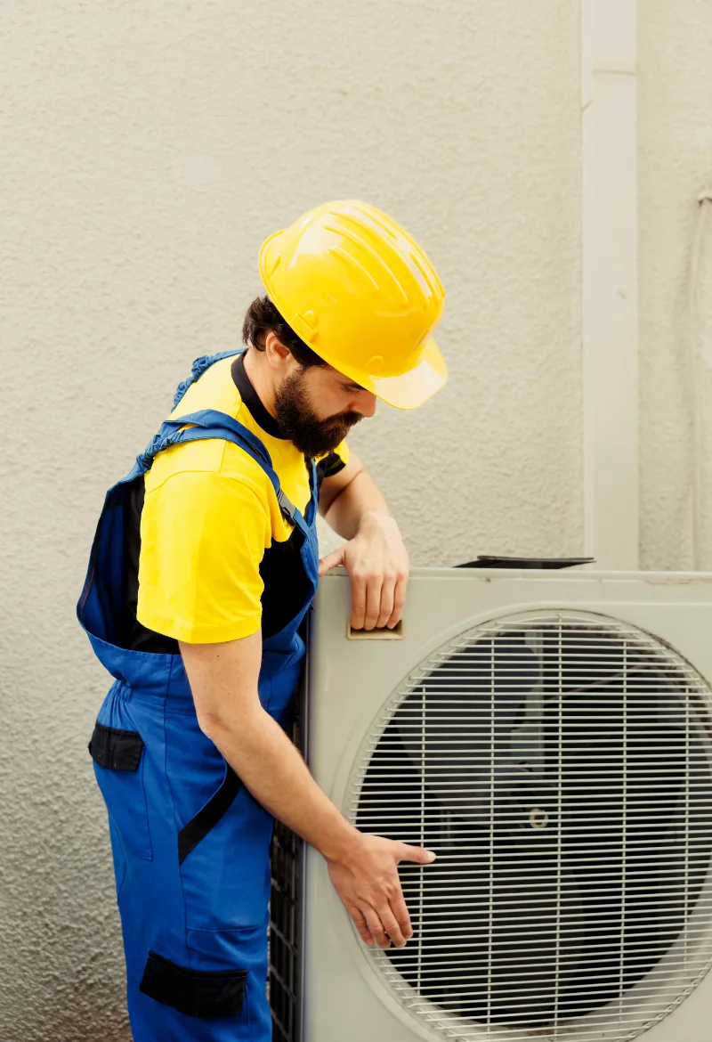 HVAC technician in a yellow helmet and blue overalls working on an outdoor air conditioning unit
