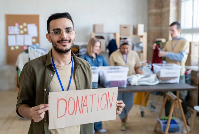 A person is holding a sign that says "DONATION" in red letters. Behind them, a group of people are working in a donation center, sorting through clothes. The setting appears to be a volunteer or non-profit organization workspace