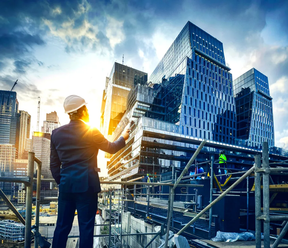 A construction manager in a suit and helmet stands on scaffolding, pointing at a modern skyscraper under construction during sunset, with workers visible in the background