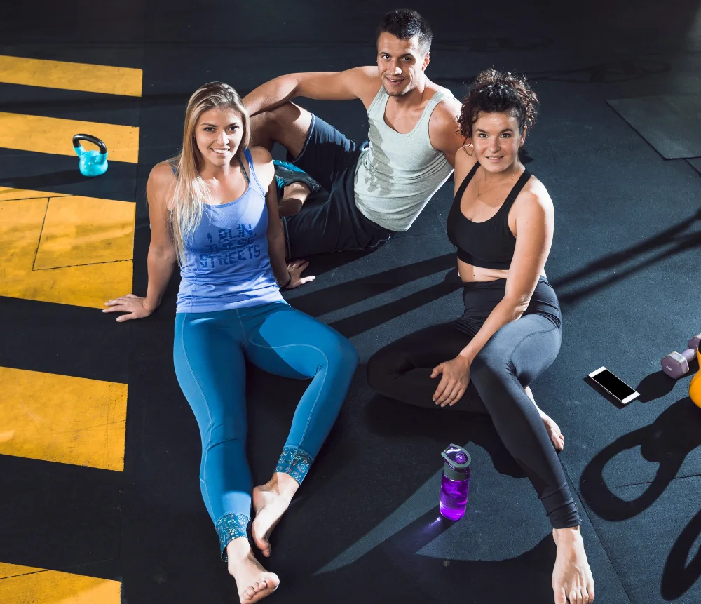 Three people relaxing in a gym after a workout. Two women and one man are sitting on the floor in fitness attire, surrounded by gym equipment such as kettlebells, dumbbells, and water bottles
