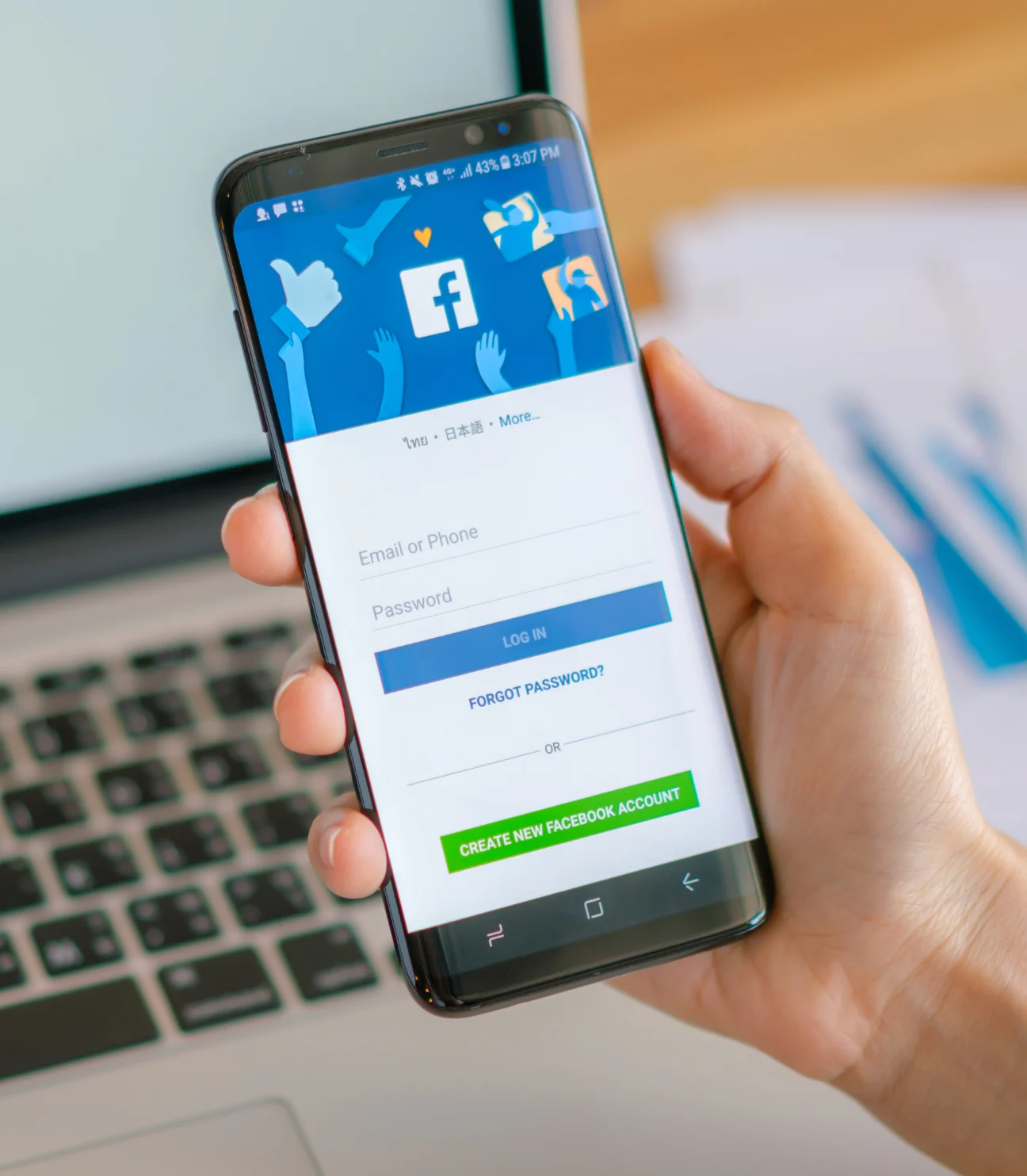 A person holding a smartphone displaying the Facebook login screen, with fields for email/phone and password. The Facebook logo and various social media icons are visible in the background, and a laptop is placed on the table in the background.