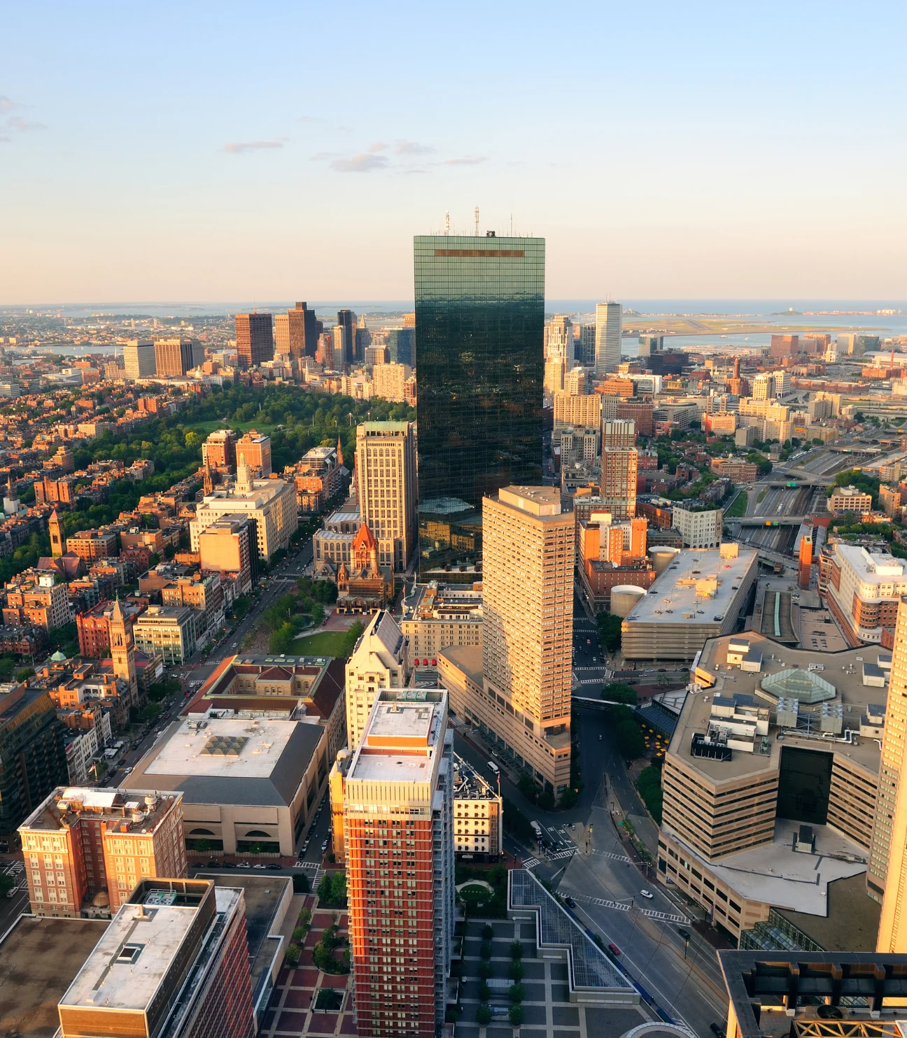 A panoramic view of Kansas City from above, showing a bustling urban landscape with tall buildings and streets. The image captures the city at sunset, with warm lighting casting over the skyline and the surrounding area, including green spaces and highways.
