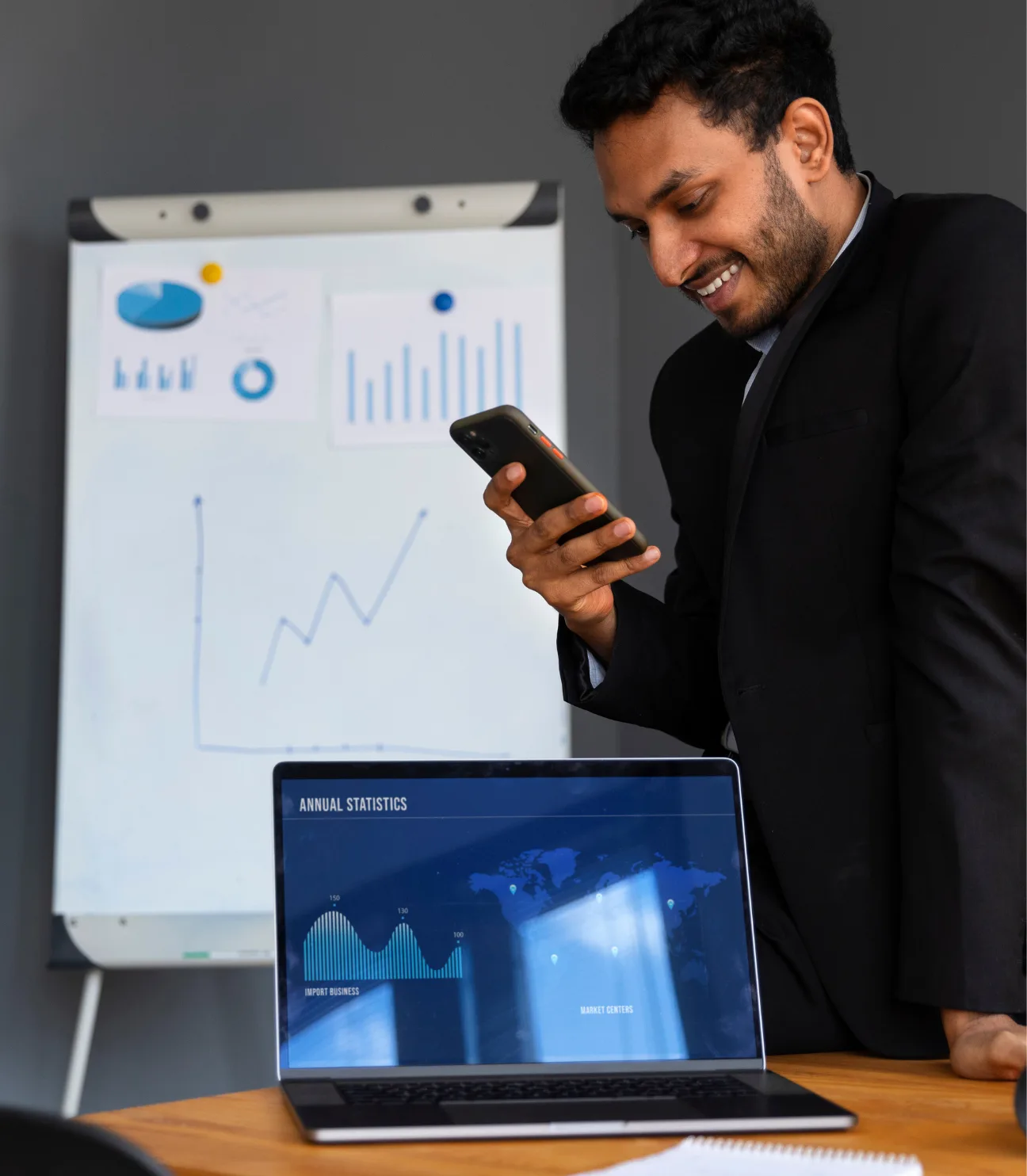 A businessman in a suit smiling while looking at his smartphone. In the background, there's a presentation board showing various graphs and charts. On the table, a laptop displays annual statistics with data on import businesses and market centers