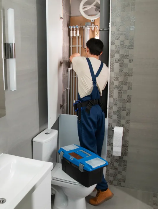 A plumber in blue overalls is working inside a bathroom, repairing pipes behind a cabinet. He is using tools and bending over a toolbox placed next to a toilet. The pipes are visible through the open cabinet door.