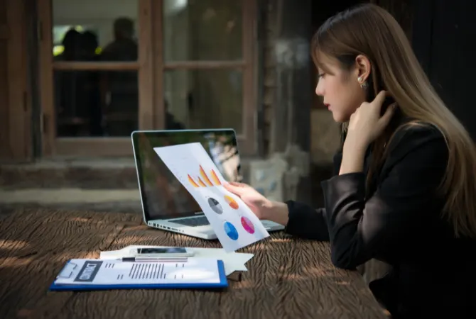 Businesswoman analyzing performance charts on paper while working on a laptop, symbolizing data-driven decisions and the results-focused approach of Pay For Performance SEO.
