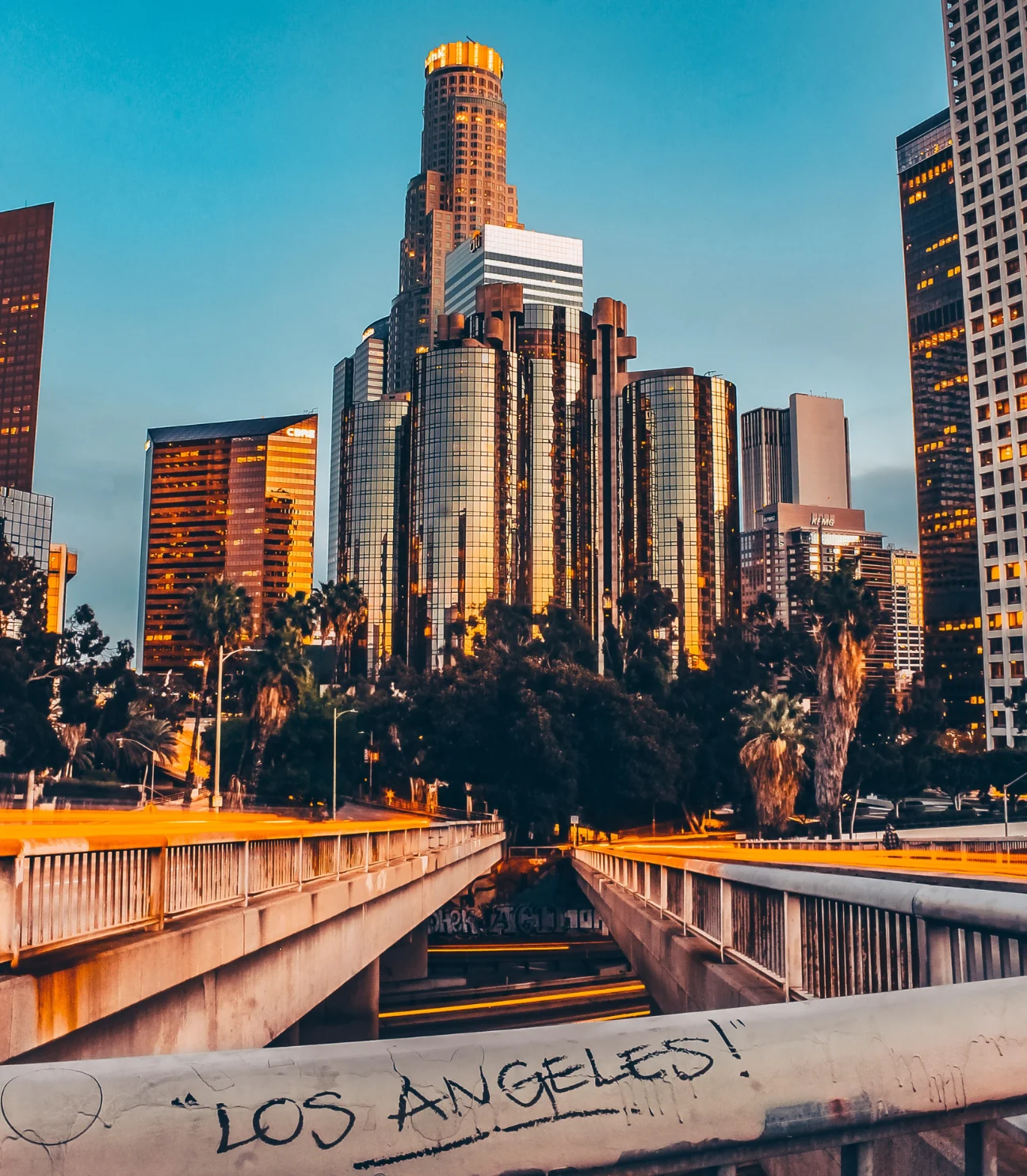 A vibrant sunset view of the Los Angeles skyline, featuring tall modern buildings with glass facades. In the foreground, a bridge leads to the city, and the words 'LOS ANGELES!' are spray-painted on the railing.