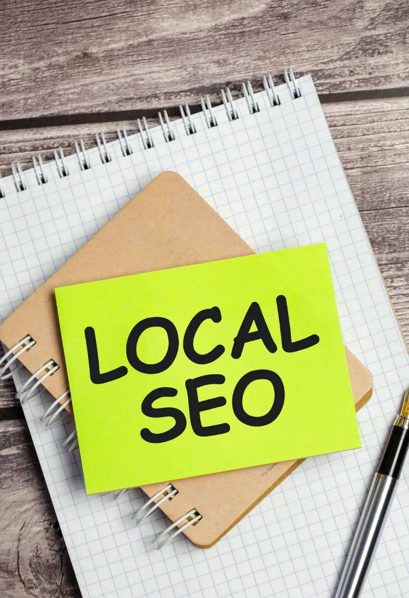 The image shows a bright yellow sticky note with the words "LOCAL SEO" written in bold black letters, placed on top of a notebook with a grid pattern and a pen beside it. This visual highlights the concept of Local SEO, which focuses on optimizing a website for local search results.