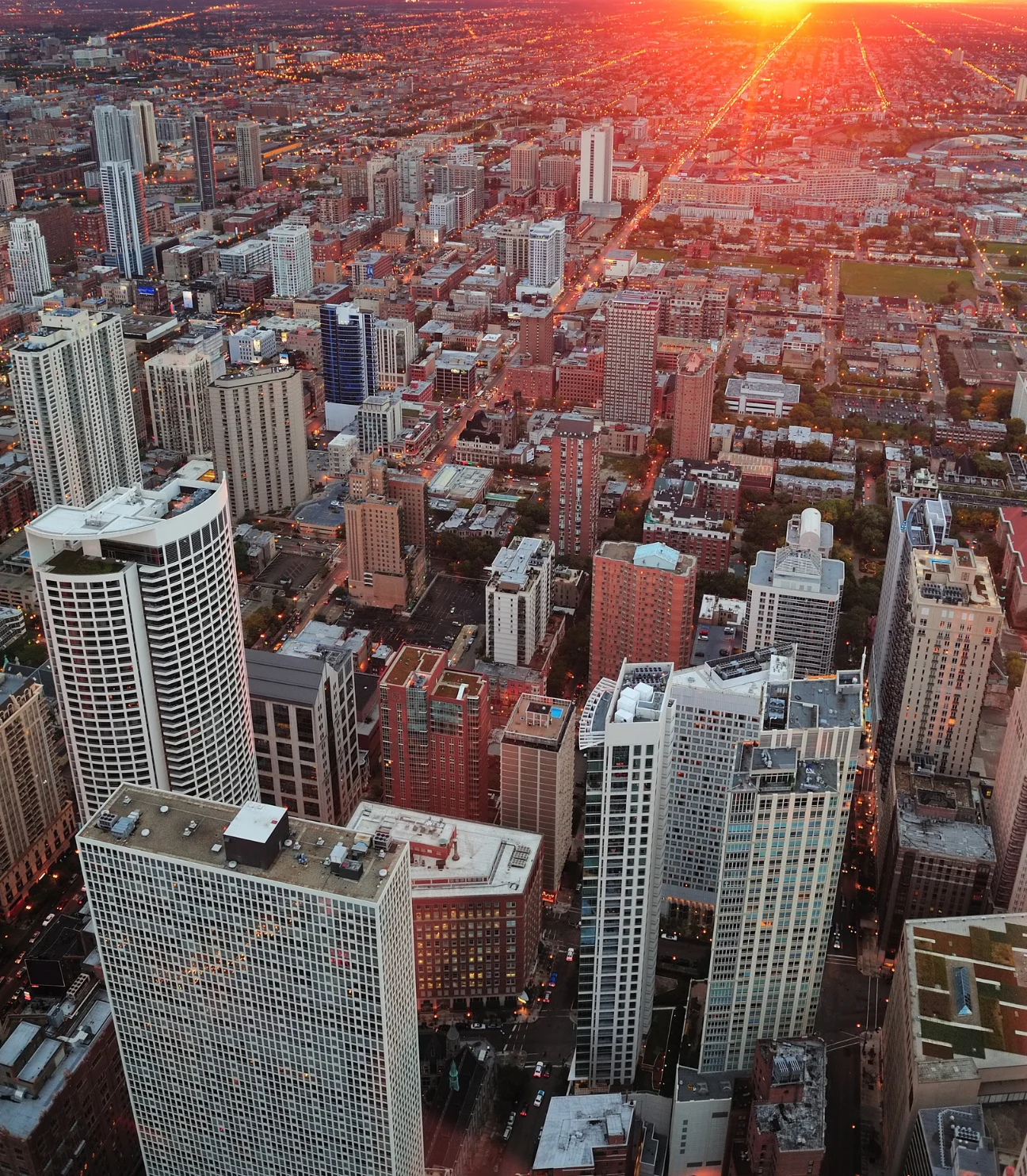 An aerial view of a cityscape at sunset, with a dense collection of high-rise buildings. The scene features city streets lit up by streetlights, and the horizon glows with the warm colors of the setting sun, casting an orange glow over the city