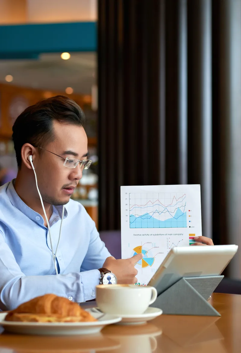 A man in a blue shirt sitting at a table, reviewing graphs and charts on a printed document while pointing at a chart. He is wearing earphones, and a tablet with financial data is on the table next to a cup of coffee and a croissant.