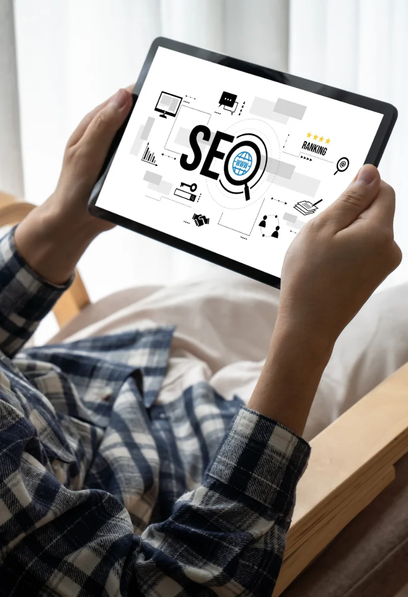 A person holding a tablet displaying an SEO concept with icons related to website ranking, keywords, and analytics. The image shows the SEO process and strategies for improving search engine visibility, set against a light background.