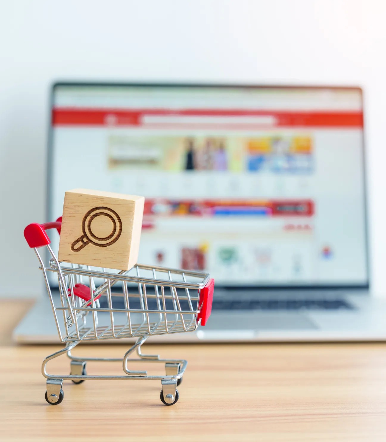 The image depicts a small shopping cart with a wooden block featuring a magnifying glass icon, placed in front of a laptop displaying an eCommerce website. This visual represents the concept of eCommerce SEO services, highlighting online shopping and search optimization.