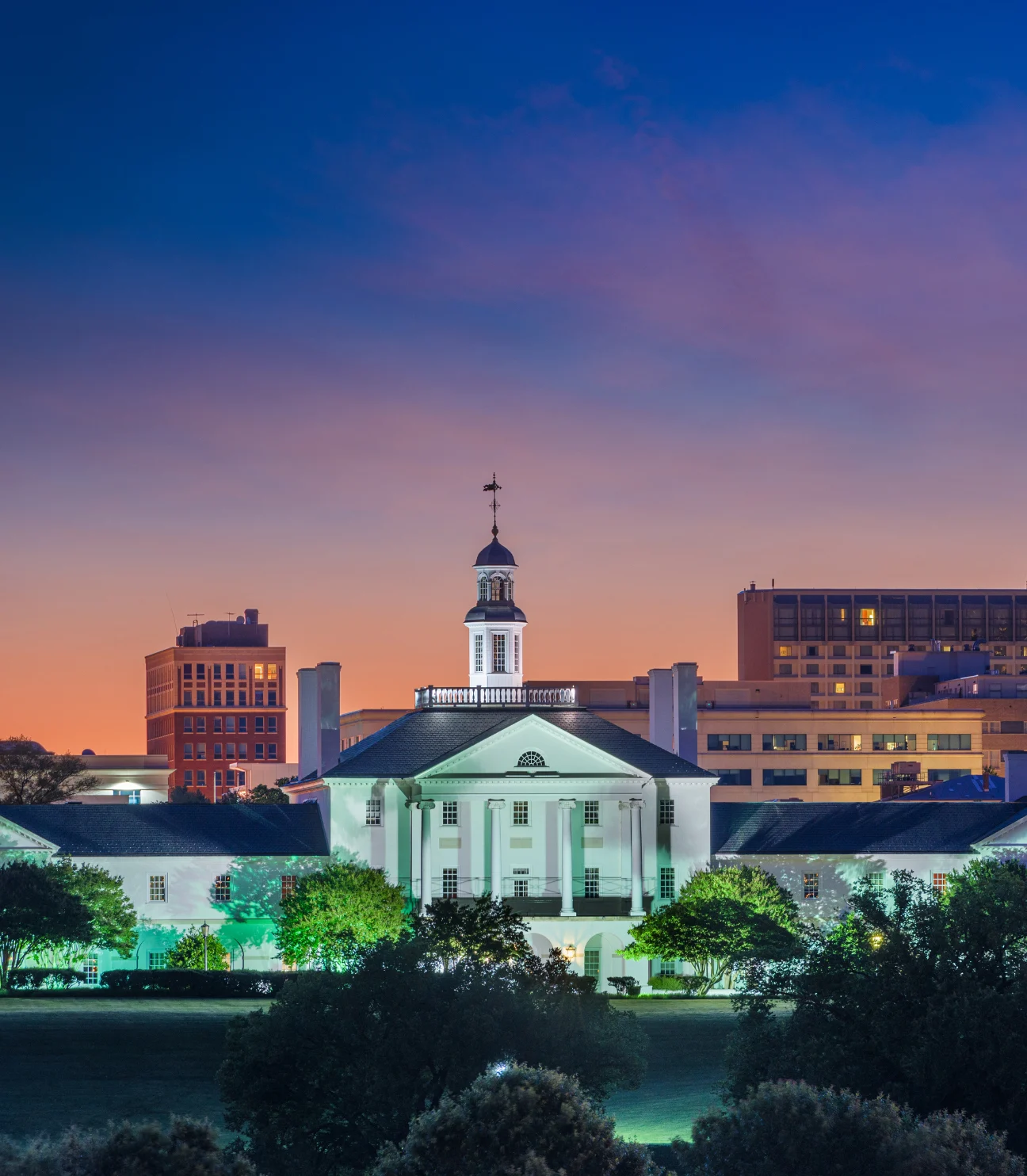 An evening cityscape showcasing a classic white building with columns, representing the essence of a prominent SEO company in Virginia. The building is lit with green lights, surrounded by trees and greenery, while a modern high-rise building stands in the background. The vibrant sunset sky adds to the scenic view, reflecting the dynamic nature of the SEO industry.