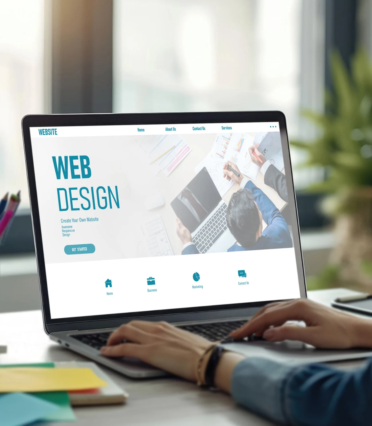 A person working on a laptop with a website design open on the screen. The website displays 'WEB DESIGN' in large letters, along with options like 'Home,' 'Business,' 'Marketing,' and 'Contact Us.' The desk is cluttered with colorful office supplies, and the image is shot with a focus on the person’s hands typing.